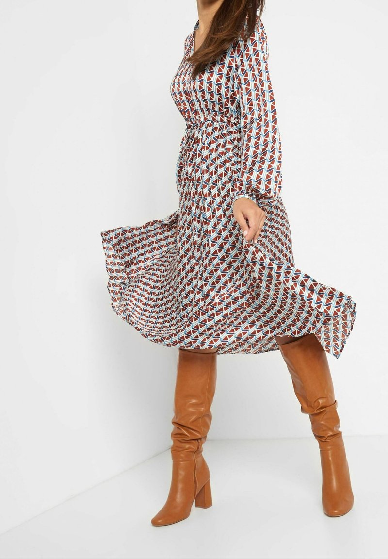 Tiskované šaty s geometrickými vzory v červené, modré a krémové barvě. Dlouhé rukávy, přiléhavý pas, splývavá sukně, kombinované s hnědými vysokými botami.