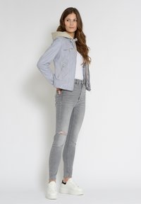 Lichtgrijze neplederen jas met een beige capuchon, met ritszakken, gecombineerd met grijze ripped jeans en witte platform sneakers.