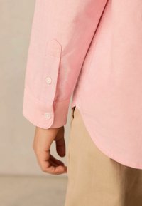 Camicia a maniche lunghe di colore rosa chiaro con polsini appuntiti e due bottoni, con un orlo curvo e tessuto morbido abbinata a pantaloni beige.