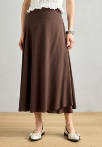 Gonna marrone alla caviglia realizzata in tessuto morbido con design svasato. Presenta una vita larga e una texture liscia, abbinata a scarpe bianche.