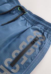 Pantaloncini sportivi blu in tessuto leggero, con tasca laterale con zip e dettagli neri, e grande stampa del logo grigia.