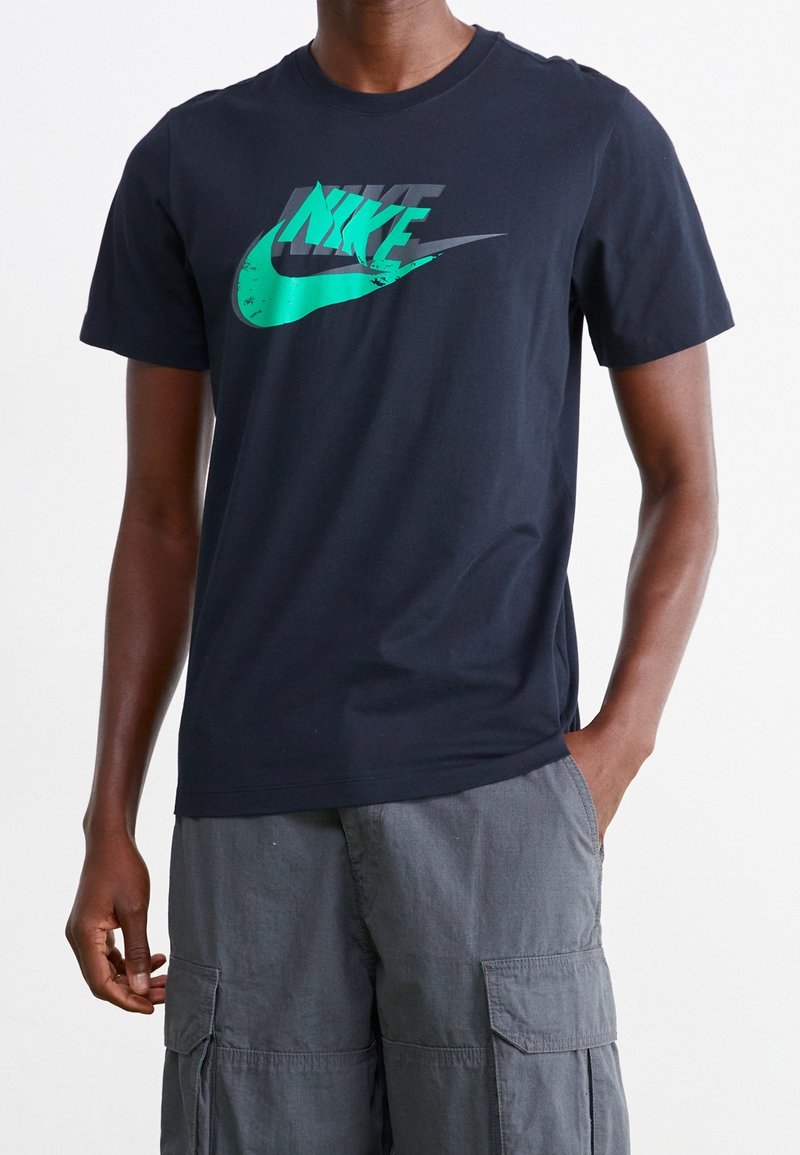 Osoba oblečená v černém tričku Nike s modrým logem a šedých cargo šortkách, stojící s jednou rukou v kapse na bílém pozadí.