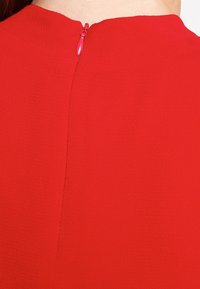 Indumento rosso con una texture liscia, collo rotondo e zip discreta sul retro. Design minimalista senza decorazioni.