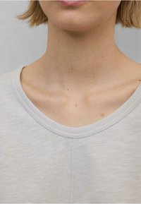 Světle šedý krátký rukáv top s širokým kulatým výstřihem. Tkanina vypadá měkce s jemnou texturou a zobrazuje delikátní švy podél výstřihu.