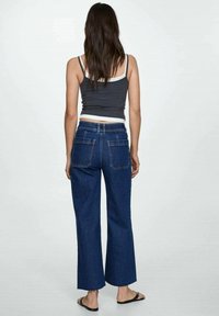 Jeans en denim foncé à jambes larges avec poches arrière, portés avec un débardeur gris. La texture est lisse avec un design classique à cinq poches.