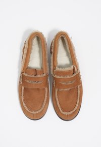 Mocasines de ante marrón con forro de shearling, correas dobles en la parte delantera y costuras blancas. Diseño de perfil bajo con suelas texturizadas.