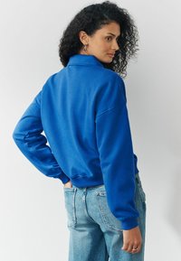 Vrouw met krullend haar in een felblauwe langlopende trui en lichtblauwe jeans, iets naar de zijkant gekeerd tegen een effen achtergrond.