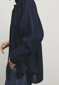 Chemise oversize bleu marine avec un col boutonné, des manches longues et une coupe décontractée, confectionnée dans un tissu texturé, portée sur un denim.