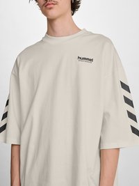 Jeune personne portant un t-shirt de sport hummel beige oversize avec des bandes chevrons noires sur les manches et un petit logo sur la poitrine.