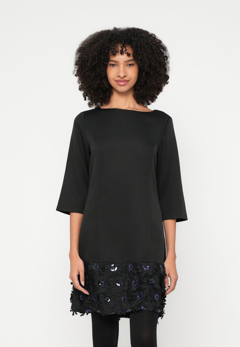 Μαύρο φόρεμα με μανίκια τρία τέταρτα, διακοσμημένο με υφασμάτινο τελείωμα με λουλουδένιες υφές, παγιέτες και πέταλα. Λείο ύφασμα και εφαρμοστή σιλουέτα.