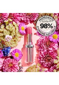 Rossetto rosa in un tubetto trasparente con accenti riflettenti, circondato da vari fiori vivaci su uno sfondo rosa. Ingredienti naturali indicati.