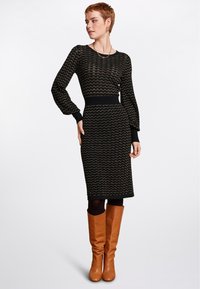 Svart stickad klänning med ett vågigt guldmönster, långa ärmar, figursydd midja och knälång fåll. Bärs med tanfärgade knähöga stövlar.