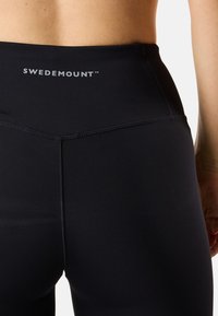 Leggings noirs taille haute avec des détails de couture et un logo SWEDEMOUNT à l'arrière. Fabriqués à partir d'un matériau extensible et lisse.