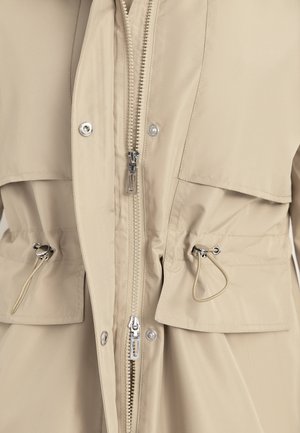 Beige Jacke, Vorderseite mit zwei Reißverschlusszügen, Druckknopftaschen mit Klappen und verstellbaren Kordelzügen mit Metallenden an der Taille.