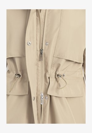 Beige jas van voren met twee ritstrekken, klepzakken met drukknopen en verstelbare tunnelsluitingen met metalen knoppen aan de taille.