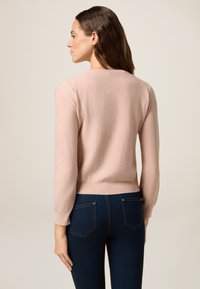 Frau mit langen braunen Haaren, die einen hellrosa Pullover mit langen Ärmel und dezenten Glitzerpartikeln sowie dunkelblauen enganliegenden Jeans trägt, die von hinten zu sehen ist.