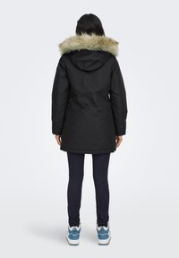 Vrouw die met haar rug naar de camera staat, gekleed in een zwarte parka met een bonttrimmed capuchon, donkere skinny jeans en wit-blauwe sneakers, tegen een effen achtergrond.