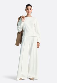 Valge liibuva lõikega dressipluus ja laia säärega püksid, mõlemad pehmest kangast. Käe peal on helepruun jakk.