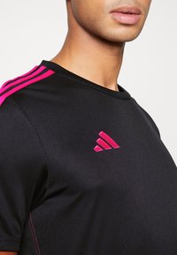 Svart idrottströja med rosa tre-stripes-design på axlarna och en matchande rosa logotyp. Slätt, lätt tyg med en enkel rund halsringning.