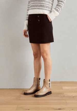 Femme portant un pull à motifs, une mini-jupe noire boutonnée et des bottines beiges, debout sur un sol en bois devant un mur gris uni.