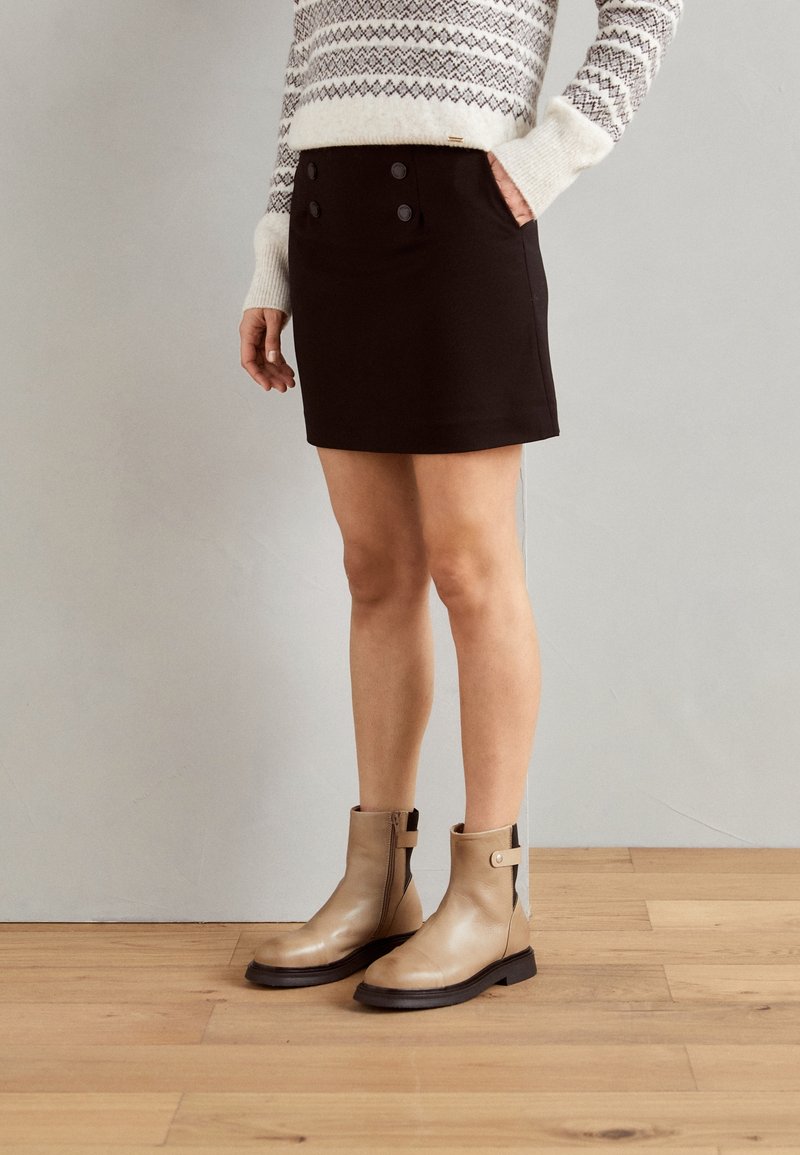 Femme portant un pull à motifs, une mini-jupe noire boutonnée et des bottines beiges, debout sur un sol en bois devant un mur gris uni.