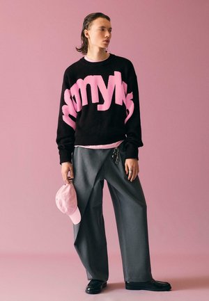 Fekete pulóvert viselő személy, nagy rózsaszín felirattal, szürke bő szárú nadrágban, fekete cipőben, rózsaszín sapkát tartva, egyszínű rózsaszín háttér előtt.