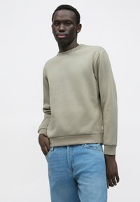 Felpa color kaki con collo tondo e maniche lunghe, realizzata in tessuto morbido; abbinata a jeans azzurri. Presenta polsini e orlo a coste.