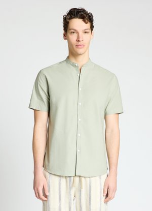 Jeune homme aux cheveux courts et bouclés, portant une chemise à manches courtes vert clair à col mandarin boutonnée et un pantalon rayé, debout devant un fond uni.
