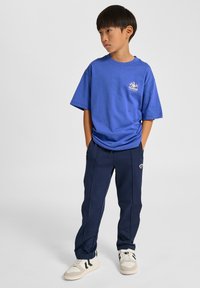 Blå bomullst-shirt med tryckt logotyp, kombinerad med marinblå sportbyxor med dragkedjor och en subtil detalj. Vita sneakers med svarta ränder.