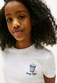 Hvid bomulds t-shirt med korte ærmer, der har et popcorngrafik i blå og røde striber, samt "Tommy Hilfiger" broderet under designet.