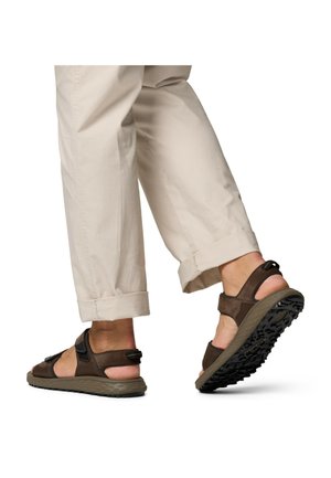 Persona con pantaloni beige risvoltati e sandali marrone scuro open-toe con suole lavorate, che cammina su uno sfondo bianco.