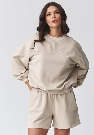 Kobieta o falowanych ciemnych włosach, ubrana w beżową, oversize'ową bluzę z napisem "MAKADAMIA" oraz dopasowane szorty, stojąca z rękami na biodrach.