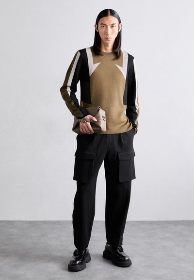 Olíva színű, kötött pulóver fekete és fehér geometrikus részletekkel, fekete cargo nadrággal párosítva. A modell egy kis bézs clutch-ot tart. Fekete cipő.