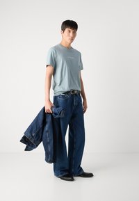 Šviesiai mėlyna T-shirt marškiniai, laisvas kirpimas, balta tekstas, derinami su plačiomis tamsiai mėlynomis džinsomis ir džinsiniu švarku, juodi batai, minimalistinis dizainas.