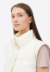 Gilet bianco imbottito con colletto alto, dotato di zip frontale e design senza maniche. Indossato sopra una camicia di colore chiaro. Tessuto liscio.
