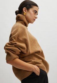 Kamel-farbener Rollkragenpullover, oversized Schnitt mit gerippten Bündchen und Saum, weicher Textur, kombiniert mit auffälligen goldenen Ohrringen.