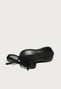 Black sunglasses with angular frames beside a black zippered case, featuring the embroidered brand name "vogue" in white.