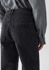 Persona che indossa jeans neri a vita alta con grandi tasche posteriori e una maglietta a maniche lunghe grigia infilata dentro, mostrata dalla vita in giù.