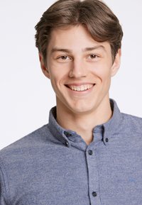 Jasnoniebieska koszula z kołnierzykiem, o miękkiej teksturze i ciemnych guzikach. Wyraziste haftowane logo na górze po lewej stronie.