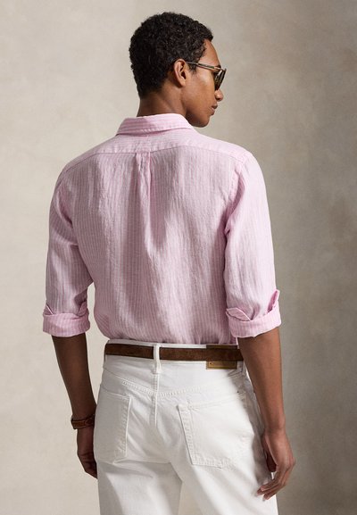 Mees, kellel on rullitud roosa triibuline särk, valged püksid, pruun vöö ja päikeseprillid, vaadates tagant neutraalse tausta vastu.