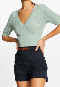 Mintgroene gebreide top met een V-hals, voorzien van een gestructureerd patroon op de mouwen. Gepaard met donkerblauwe gestreepte shorts en een leren bruine riem.