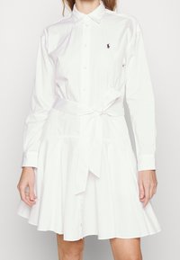 Biała bawełniana sukienka koszulowa z kołnierzykiem, długimi rękawami, guzikami z przodu, związanym pasem w talii oraz rozkloszowaną spódnicą z fałdami.