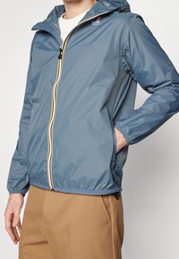 Blaue Zip-Jacke aus leichtem, wasserabweisendem Material. Verfügt über eine Kapuze, elastische Bündchen und kontrastierende Reißverschlussdetails in Gelb und Weiß.