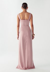 Moteris su surištais plaukais, apsirengusi be rankovių, iki grindų siekiančia, švelniai rausva satino suknele, su auksine apyranke ir kabančiomis auskarais, stovi prieš baltą foną.