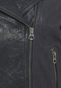 Närbild av en svart läderjacka med silverfärgad dragkedjor, en delvis stängd och en på ett litet ficka, som visar textur och sömnadsdetaljer.