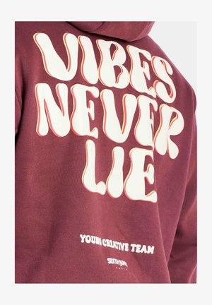 Bordeauxfarbener Hoodie mit auffälliger, retro inspirierter, weißer Schrift: "VIBES NEVER LIE" und "YOUNG CREATIVE TEAM" nahe dem Saum. Weicher Stoff.