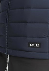 Gros plan sur une veste matelassée bleu marine avec fermeture éclair frontale et étiquette de marque rectangulaire noire AIGLE 1853 près de l'ourlet.