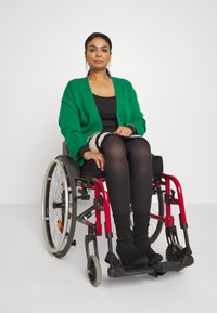 Groene oversized cardigan over een zwarte top, zittend in een rode rolstoel. Zwarte panty en kniehoge zwarte laarzen. Eenvoudige achtergrond.