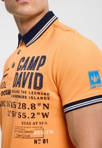 Orange polo shirt with a navy striped collar, featuring navy embroidery and printed graphics, including coordinates and a logo patch on the sleeve.