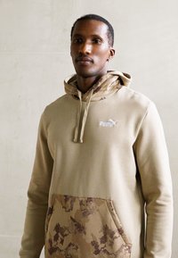 Joven vestido con un sudadera beige de Puma con bolsillo y capucha de estampado de camuflaje, de pie contra una pared lisa de color claro.